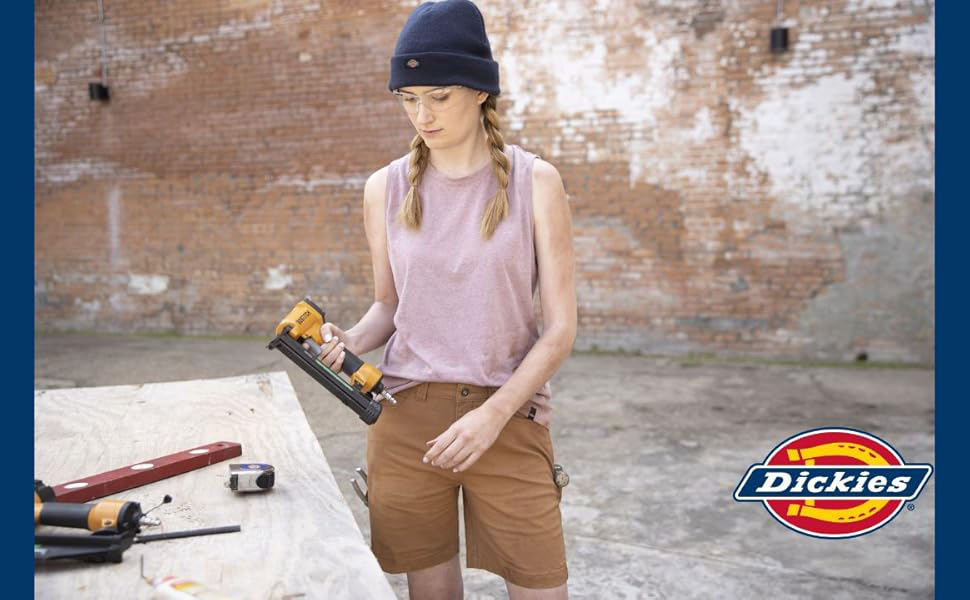 Woman in hat and shorts holding nail gun in front of brick wall